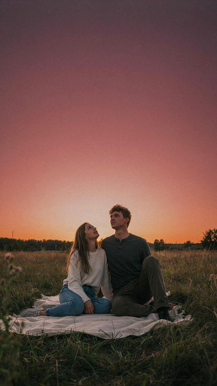Two people sit on a blanket, watching the colorful sky change at sunset.