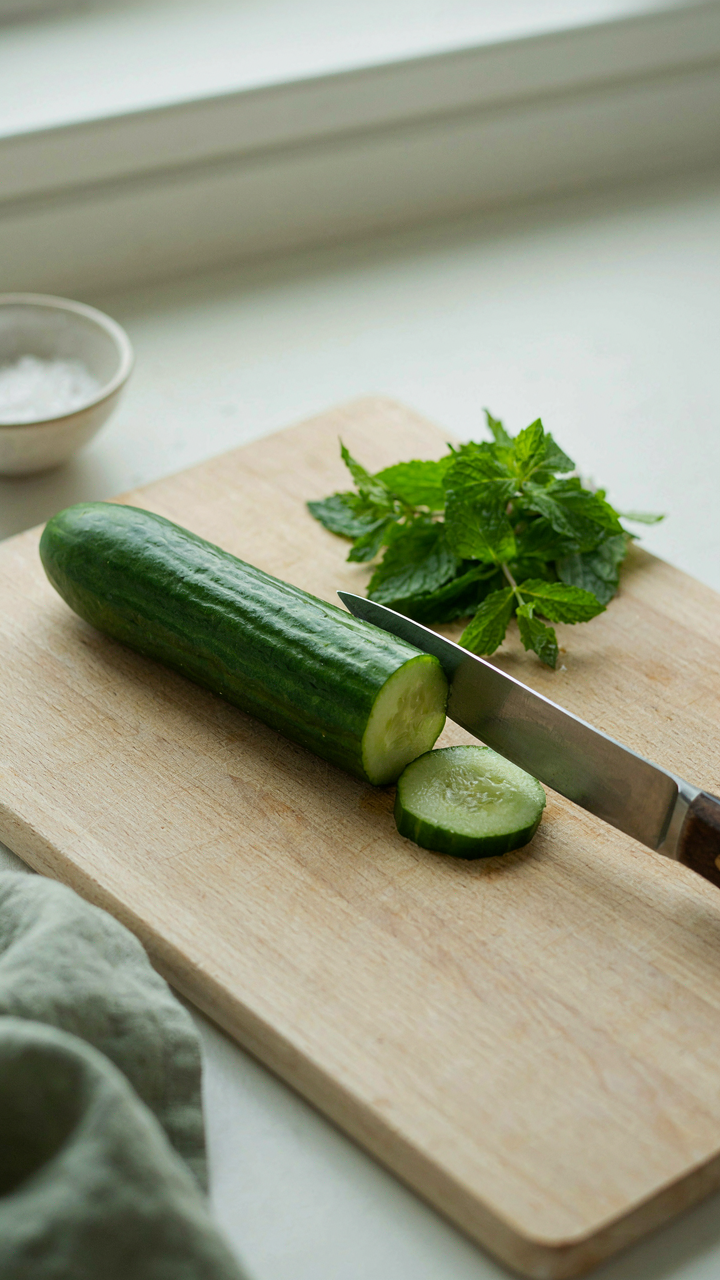 A ripe cucumber is carefully sliced by hand on a wooden cutting board with fresh mint.