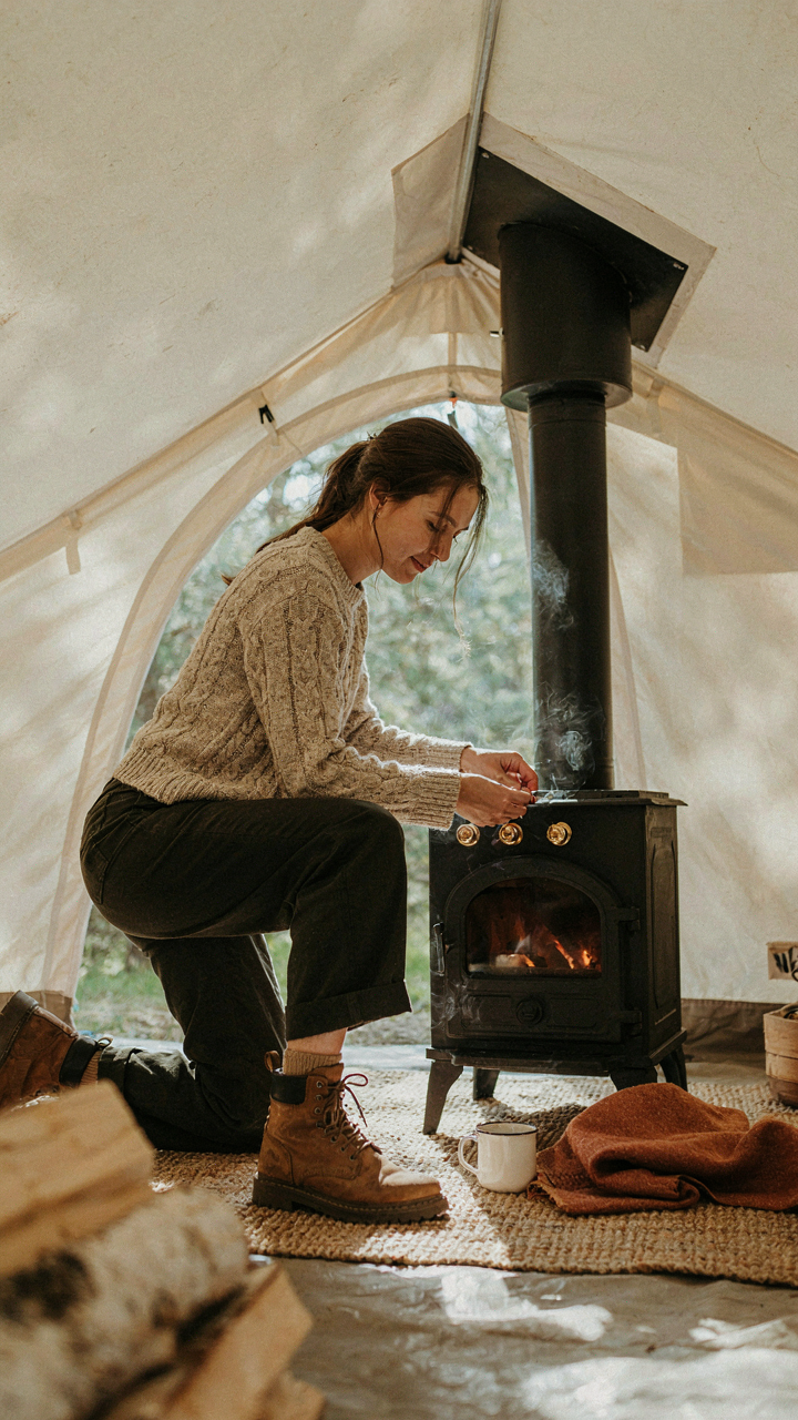 A person adjusts a wood stove inside a glamping tent.