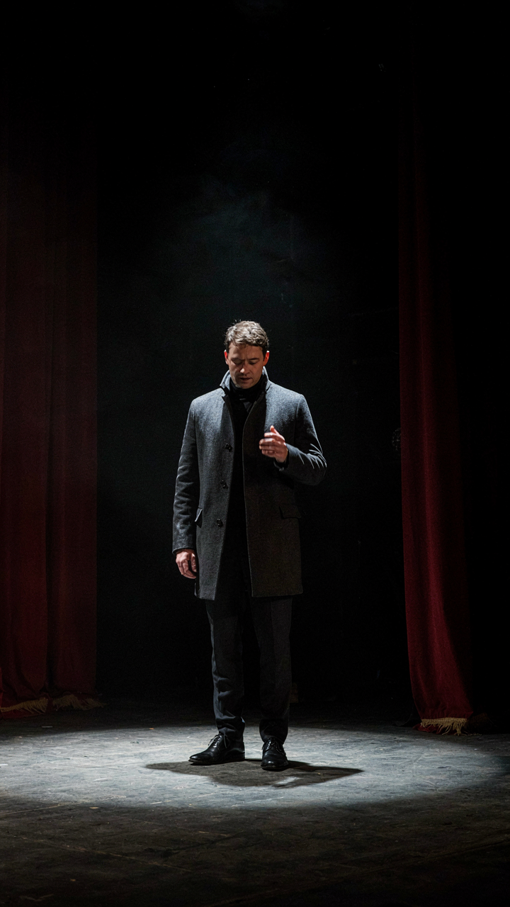 An actor on a stage lit by a single spotlight, surrounded by deep shadows.
