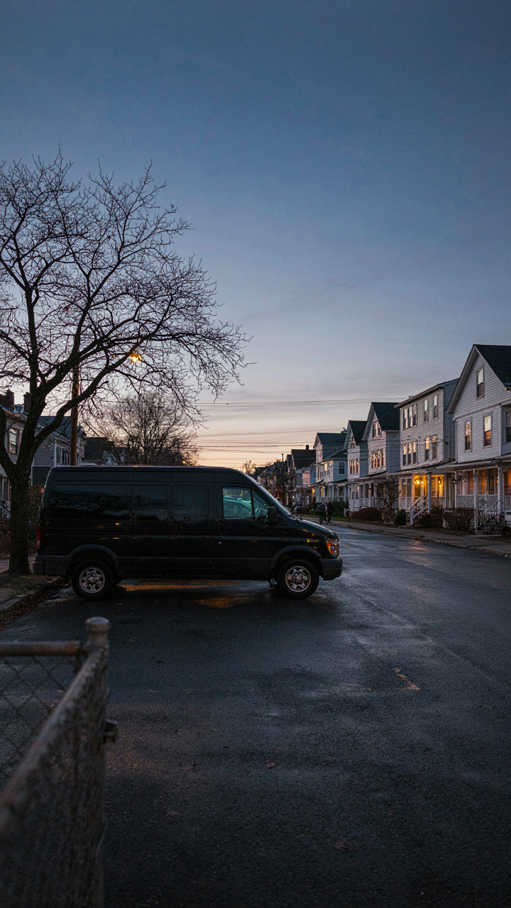 A dark, unmarked van is parked discreetly on a quiet residential street at dusk.