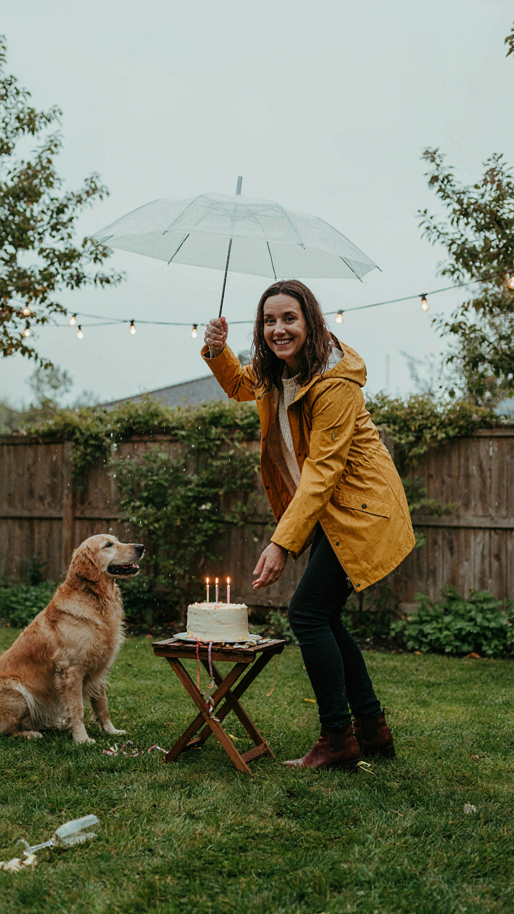 An adult smiles, holding an umbrella over a cake as rain starts.