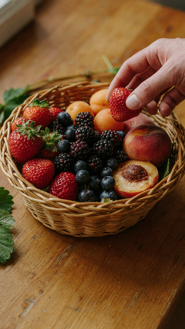 A hand selecting fresh, ripe berries and fruits from a wicker basket.