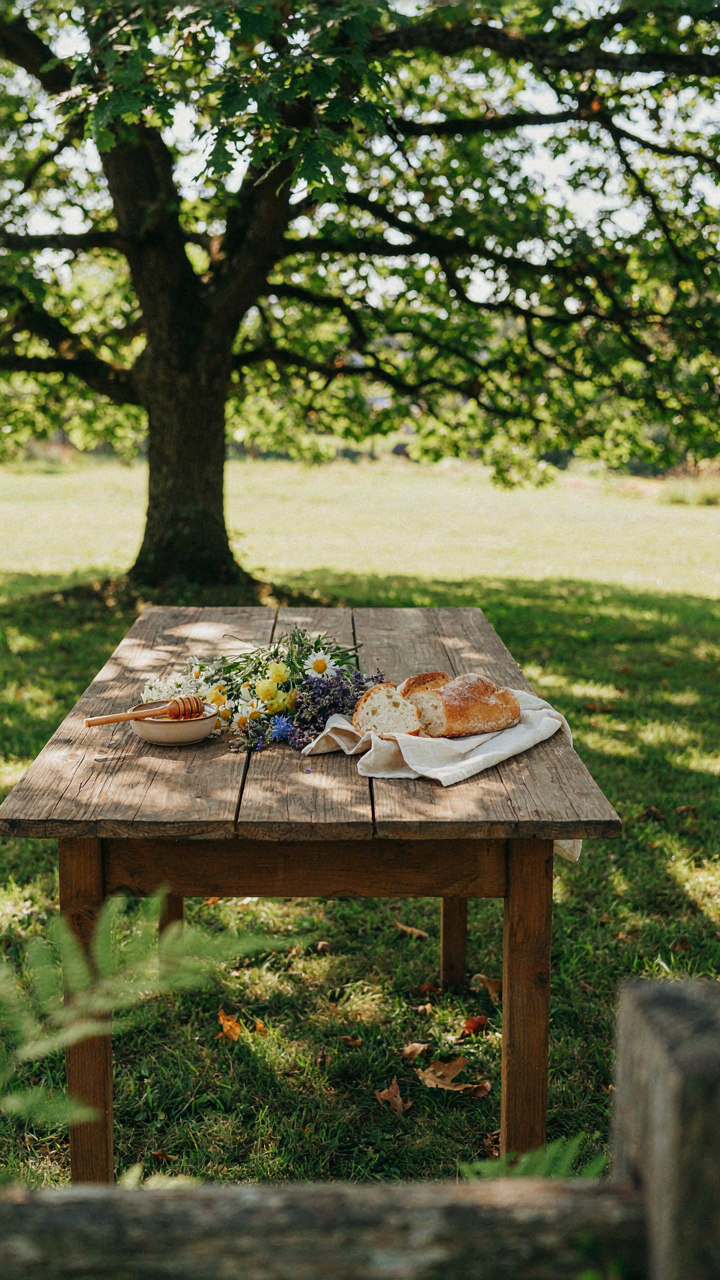 A rustic wooden table under an oak tree has wildflowers and natural elements.