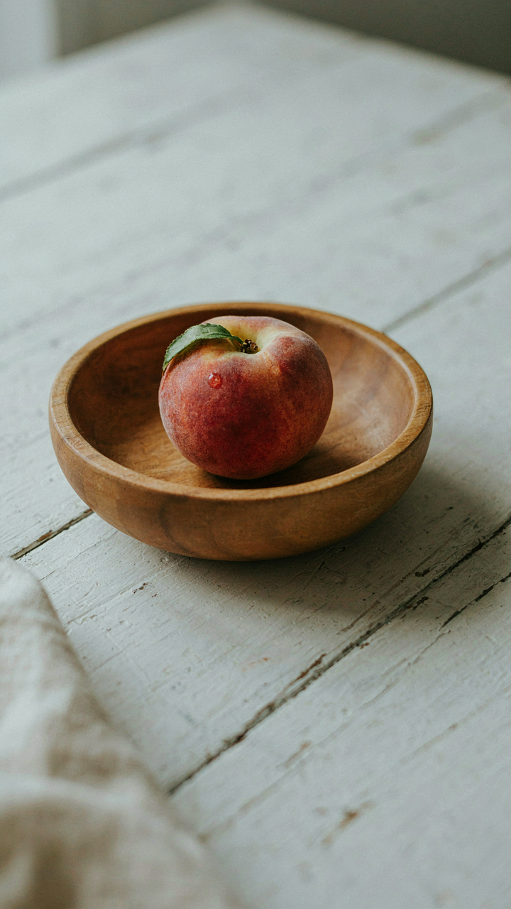 A single, perfect peach in a wooden bowl on a white table.