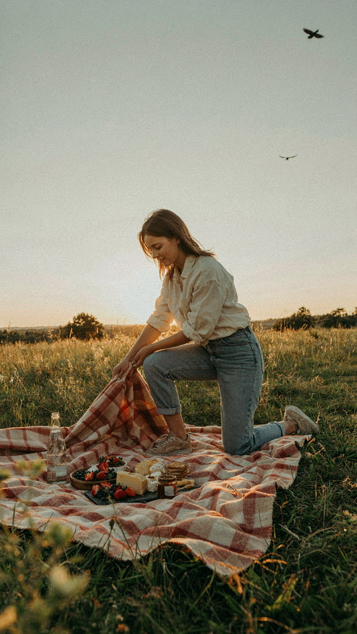 A person spontaneously spreads snacks and a blanket on the grass at sunset.