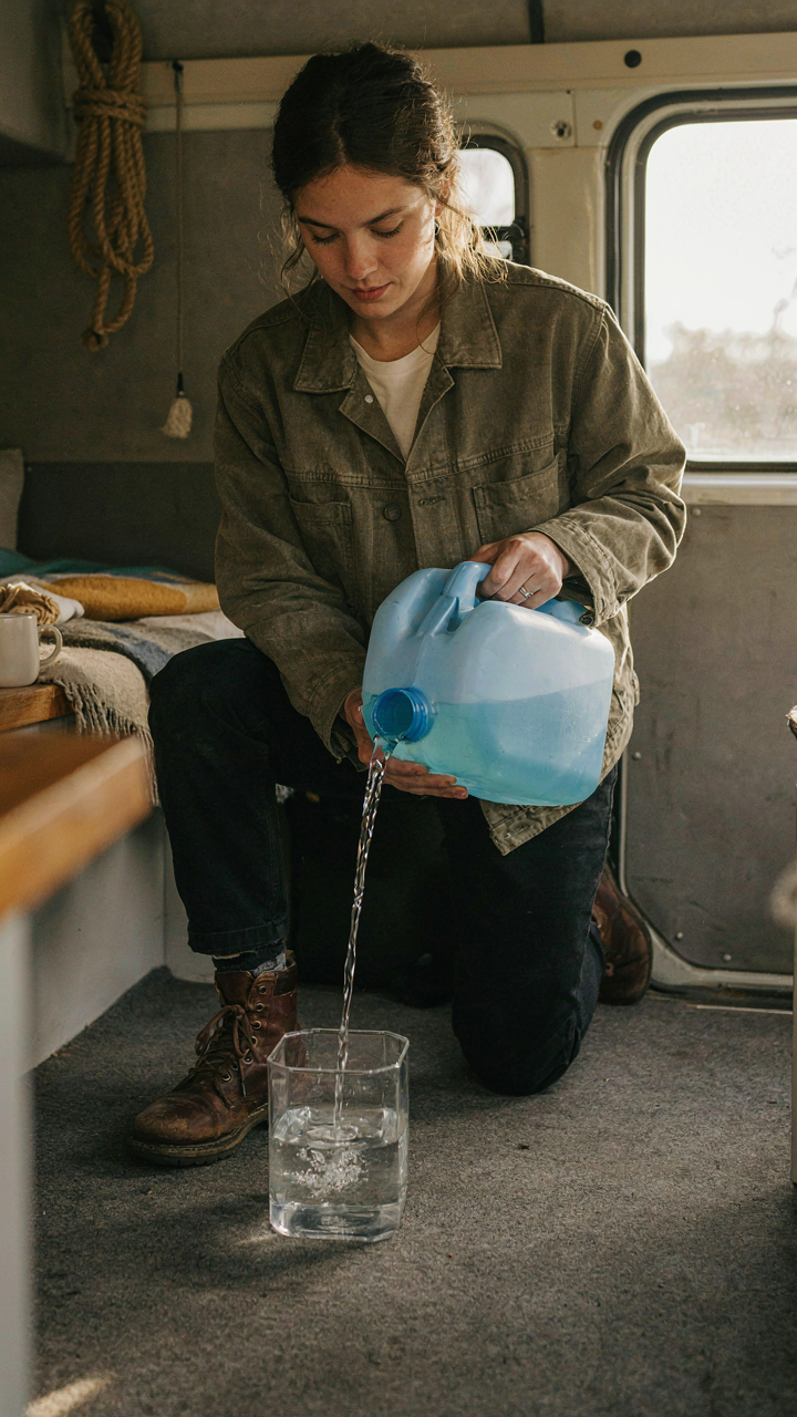 A person carefully fills a small water tank from a larger one in a van.
